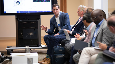 A group of people seated in a semi-circle in an office or conference room, engaged in a discussion. One person is speaking and gesturing with their hand while others listen. A large screen in the background displays presentation content.